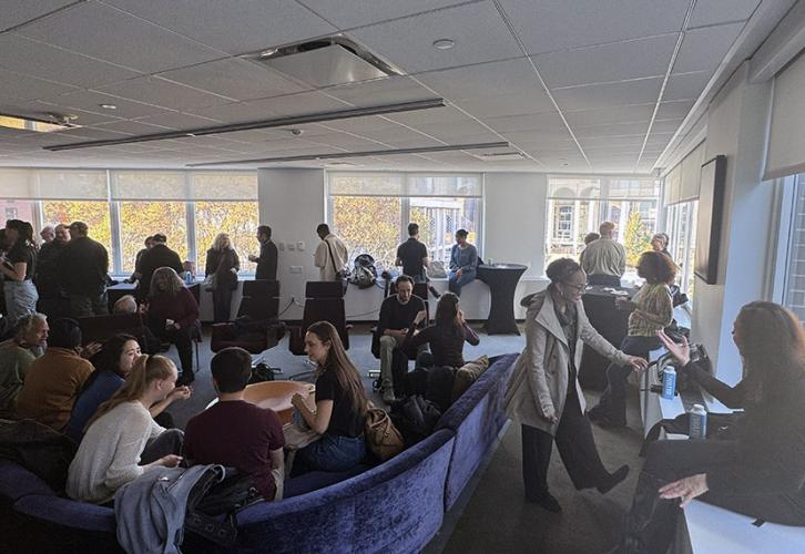 Many people are sitting on a purple velvet couch. Other people are standing around talking and drinking coffee.