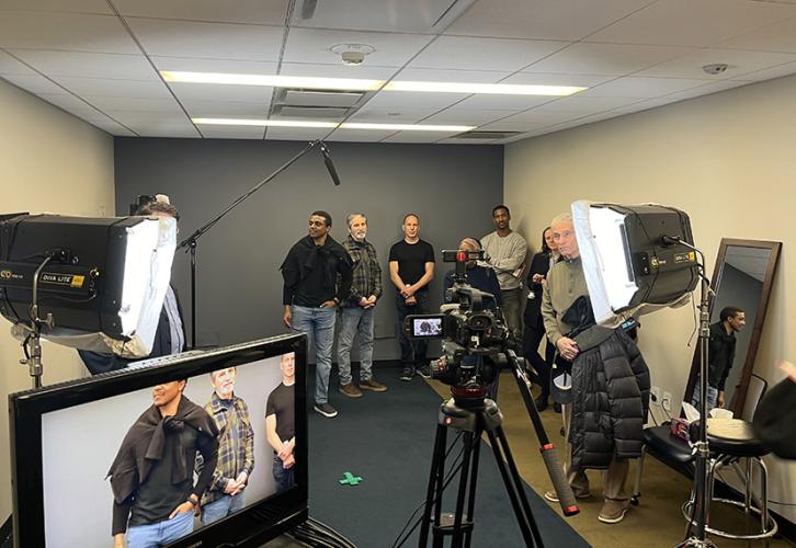 A group of people stand in a studio in front of a dark gray wall with camera equipment around them.