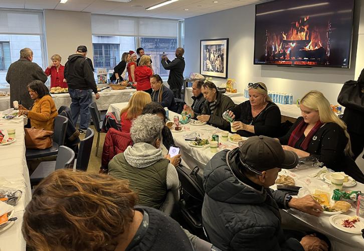 A large, diverse group of people are seated in rows at tables inside a room. There is a group of members standing in the back capturing a small group photo. A yule log video appears on the screen in the background.