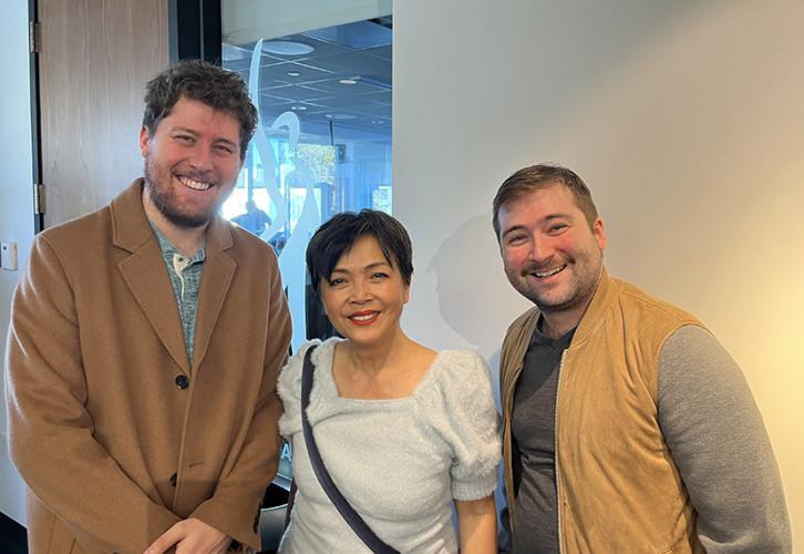 A man in a brown jacket, a woman in a white top and another man in a tan vest stand in front of a wall in an office. They are smiling.