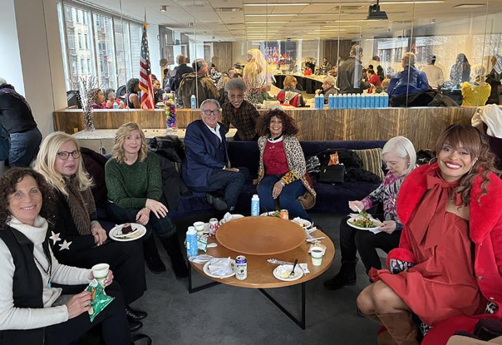 A diverse group of eight people are seated wearing festive attire. Behind the group is a view of the Ken Howard Boardroom with members seated enjoying the event.