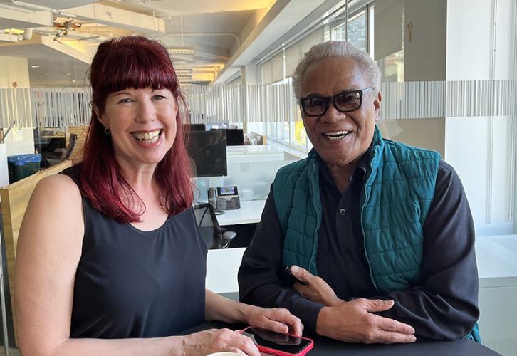 A woman wearing black and a man in a teal vest and glasses sit at a table in an office. Both are smiling.