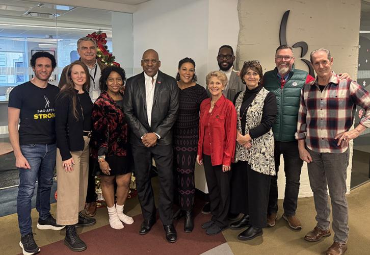 A diverse group of eleven people stand facing forward in an office. A Christmas tree is visible behind them.