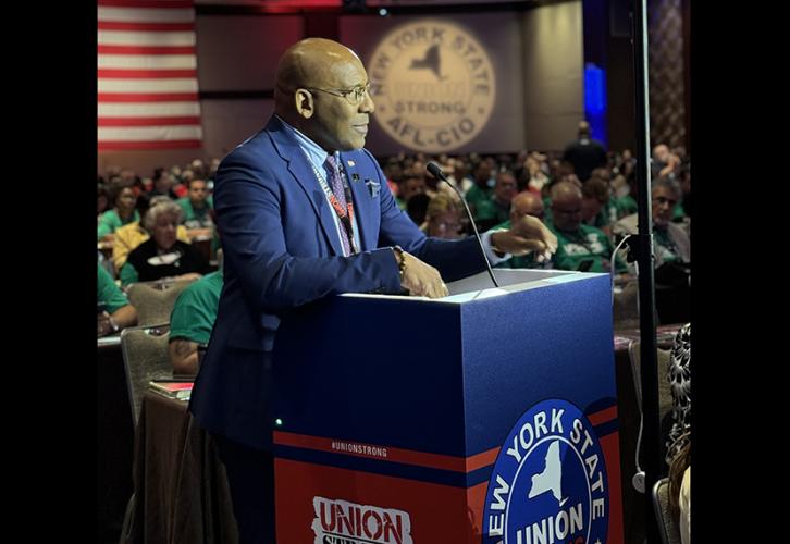 Ezra Knight in a navy suit stands behind a red, white and blue podium which reads Union Strong, New York State AFL-CIO and has the outline of the state of New York in white.