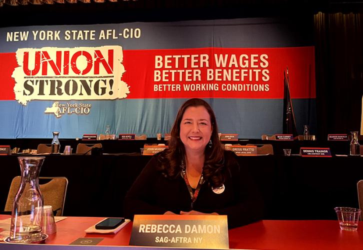 Rebecca Damon in a black blazer sits at a dais in front of the stage. A red banner stretches across the stage that reads “Constitutional Convention 2024. Better Wages, Better Benefits, Better Working Conditions. New York State AFL-CIO: Union Strong” in white text.