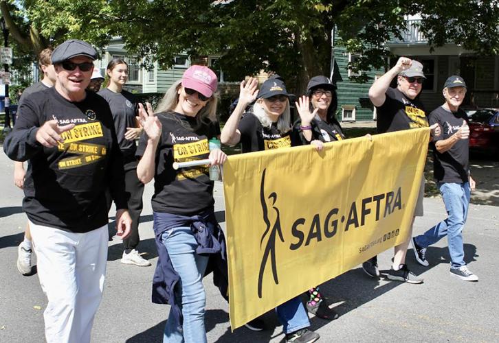 Six people carry a SAG-AFTRA parade banner with black text on a yellow background. They are walking in the street and some have fists raised. 