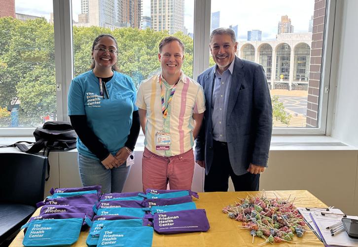 Three people stand behind a table with a yellow tablecloth. On the table are purple and blue pouches and a pile of lollipops. Behind them, Lincoln Center is visible through the window. 