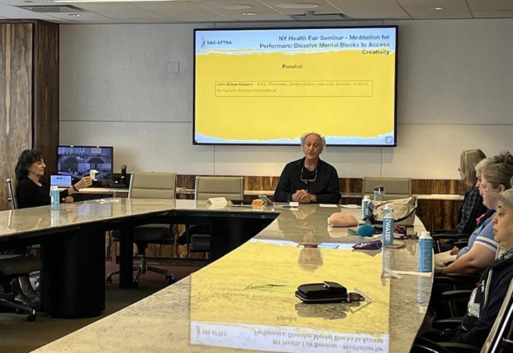 An instructor sits at the head of the table with additional people seated to the sides of the table. Behind him on a screen, in black text on a white and yellow background is the name of the presenter, John Dileva Halpern.