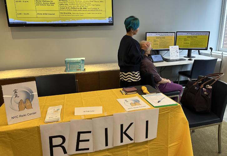 Behind a table with a yellow tablecloth and a sign that says REIKI in black text on a white background is a person seated in a chair, while another person stands behind the chair performing reiki.