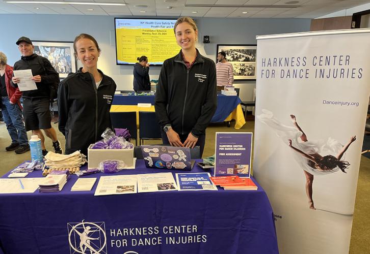 Two people stand behind a table with a purple tablecloth with white text that reads Harness Center for Dance Injuries, NYU Langone Health. Beside them is a pull-up banner with a dancer on a white background and purple text that reads Harkness Center for Dance Injuries.