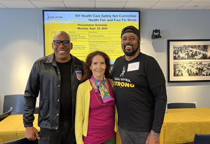 A group of three people stand in a row. The people on the ends wear black clothing. The person in the center wears a yellow sweater and colorful scarf over a pink shirt. The person on the right wears a T-shirt that reads SAG-AFTRA Strong in white and yellow text. Behind them is a screen with black text on a yellow and black background showing the schedule of seminars taking place at the health fair.