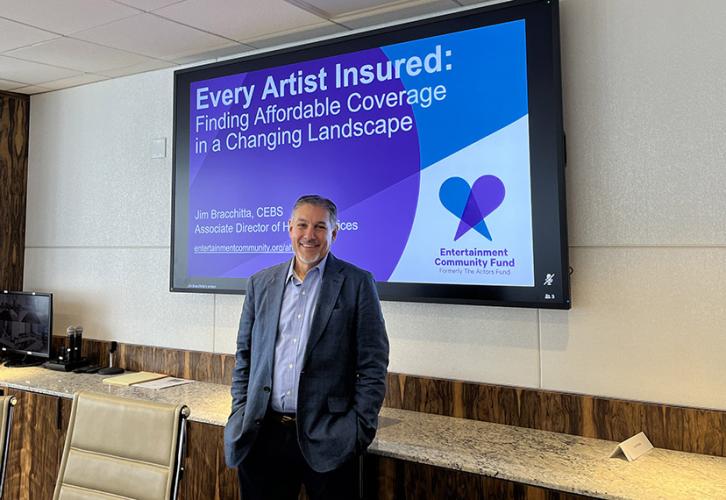 A person stands in front of a screen with white text on a purple, blue and white background that reads, “Every Artist Insured: Finding Affordable Coverage in a Changing Landscape” with an Entertainment Community Fund logo in the lower right corner. 