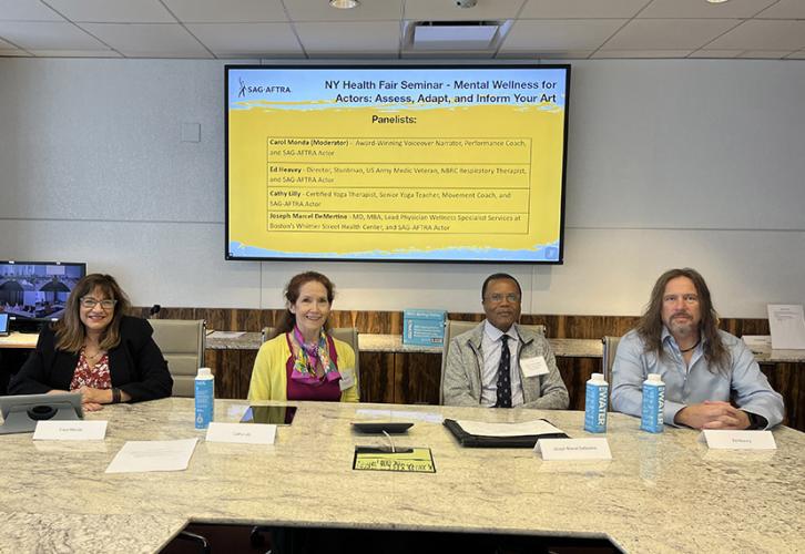 Four people sit at a table. Behind them on a screen in black text on a yellow and white background their names are listed, along with the event title. 