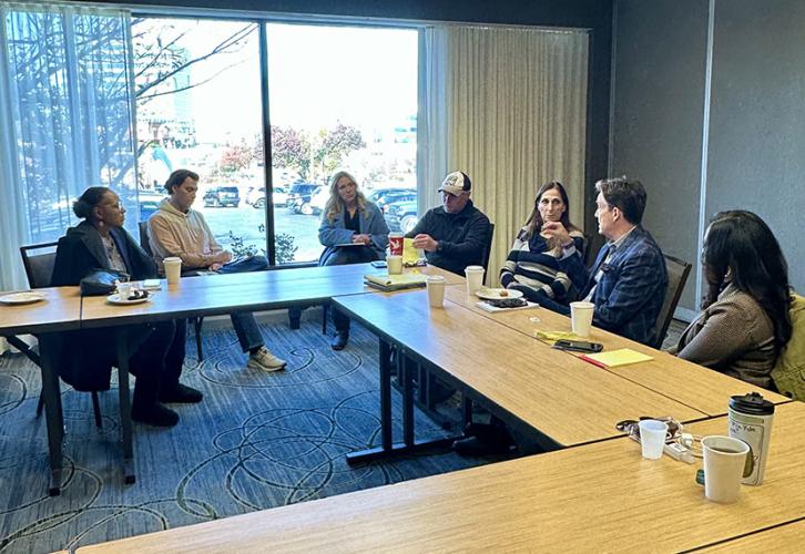 A diverse group of people have a discussion around a U shaped wooden conference table.