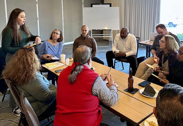 A diverse group of people listen as a person facilitates a discussion in a hotel conference room. All are seated except one woman, who is standing. There is an easel in the background.