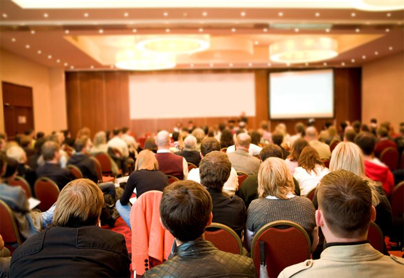 A large group of people sitting in chairs in a large conference room, their backs facing the viewer. In the front are two projectors and a blurry speakers' table.