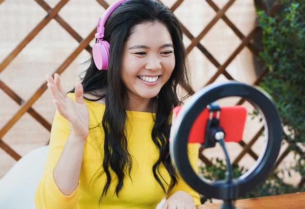 Image of female in yellow top wearing headphones looking at mobile with ring light behind.