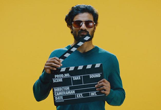 Image of a gentlemen holding a clapperboard wearing a green long sleeve and wearing dark shades standing in front of a yellow background.