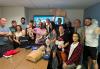 A diverse group of 16 people stand and kneel in two rows for a group photo. Many are smiling as they hold various pieces of CPR practice equipment, including two infant CPR mannequins and two adult CPR mannequins, one of which is dressed in a black SAG-AFTRA T-shirt. Horrigan, back row, holds an adult CPR mannequin above his head.