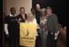 A diverse group of men and women stand for a photo against a gray wall. A women, center, holds up a small yelllow banner bearing the SAG-AFTRA logo.