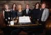 Six individuals--four females and two males--stand at a table where a cake is in the center. 