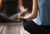 Female seated cross-legged in a blue tank top and dark grey yoga pants with the back of her right hand resting on her right knee. Pinky, ring and middle finger pointing forward with her index and thumb touching to form a circle