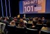 An audience seated in a theater. The setting is a dimly lit auditorium with vertical blue light strips illuminating the walls. On the stage at the front of the room, two presenters stand under a large screen displaying a slide that reads "SAG-AFTRA 101" with an image of a clapperboard. One presenter stands at a podium, while the other is speaking. The audience includes individuals of various ages, attentively facing the presenters.  