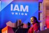 Powell stands behind a podium on stage. She wears a pink blazer. Her gaze looks out into the crowd. Behind her is a large banner with “IAM Union” in all-white, capitalized lettering with “Union” in a smaller, widely-spaced font.