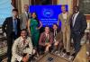 A diverse group of eight pose in an ornately decorated dining room. All are in business attire. Broussard, Talbert and Raskind crouch on the ground. Behind them on a flatscreen is an image of the White House logo and “White House Creator Economy Conference” in white text.