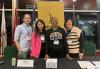 A man and three women, dressed casually, stand behind a black table. They wear lanyards around their necks and there is a yellow SAG-AFTRA banner behind them, aslong with four flags.