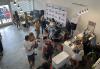 A group of diverse actors and Hawaii International Film Festival staff mingle and eat pupus in an artsy front lobby of the HIFF offices. There is a large cooler and staff working on laptops helping members purchase film festival tickets. The floor is unfinished concrete and the cooler and tables are stark white. On the right side of the space there is a step-and-repeat set up with three directors chairs placed in front for posed pictures.