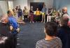 A large group of diverse people gather in a circle in the SAG-AFTRA New England Local’s conference room, one member holding a yellow ball while they prepare for an acting exercise.