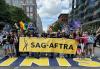 A large diverse group of people wearing and holding LGBTQIA+ SAG-AFTRA pride T-shirts and signs march on Boylston Street and cross over the finish line of the Boston Marathon as they participate in the Boston Pride Prade. People in the middle of the group hold a long, gold horizontal banner that reads “SAG-AFTRA.”