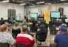 A speaker stands at the front of a spacious well-lit room, gesturing while addressing a diverse group of people seated in rows. Behind the speaker, a screen displays "Film Louisiana" alongside a yellow SAG-AFTRA banner. People seated in chairs listen.