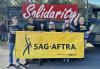 Six people stand along the side of a truck in front of a sign that says “Solidarity” in white text on a red background. They are holding a SAG-AFTRA parade banner with black text on a yellow background.