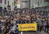 A large group of SAG-AFTRA members are gathered on a street. They are wearing black T-shirts with white and yellow text that reads “SAG-AFTRA on Strike.” They hold signs that read “SAG-AFTRA Video Game Strike” in white and yellow text on a black and purple background with the image of a fist holding a video game controller. The people at the front of the group hold a SAG-AFTRA parade banner with black text on a yellow background.