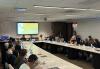 People sit around a large boardroom table. At the front, one person in a red shirt and gray jacket stands and addresses the attendees. Behind them on the screen is a slide that reads “Field Services Department Presentation with Jim Damis, Executive Director, Field Services” in black text on a yellow and white background.