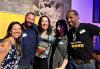 A diverse group of mixer attendees stand together and smile in front of a yellow banner in a room with windows and art hanging on the wall.